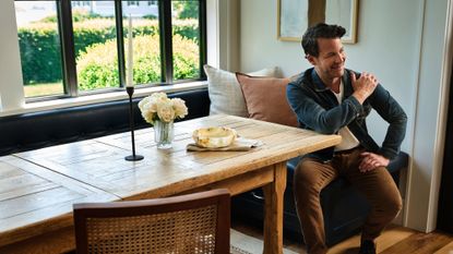Nate Berkus poses alongside his Rugs USA collaboration inside of a sunlit breakfast nook with a wooden table