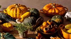 A selection of Le Creuset pumpkin Dutch ovens on a wooden kitchen table surrounded by plates of gourds. 