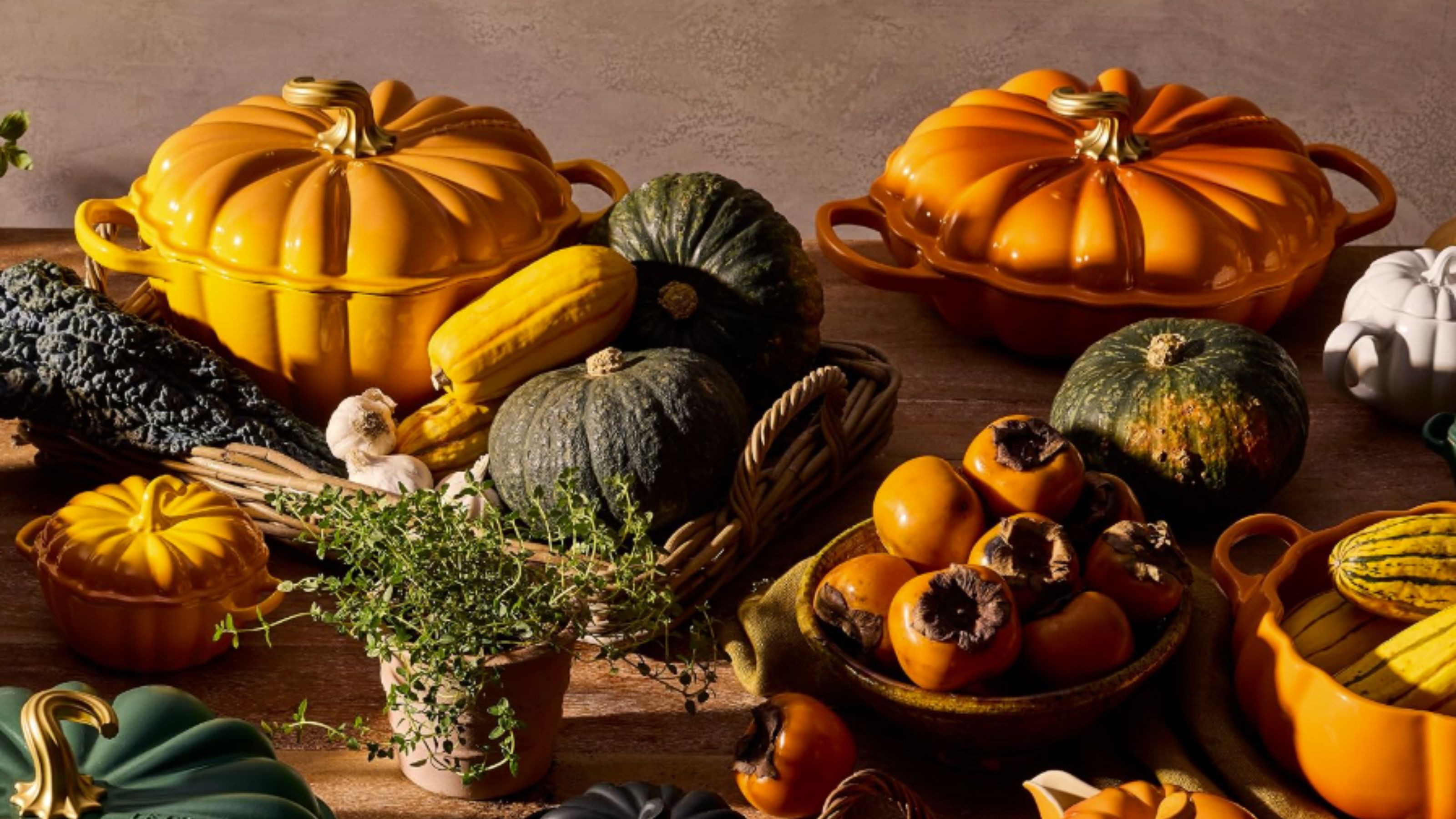 A selection of Le Creuset pumpkin Dutch ovens on a wooden kitchen table surrounded by plates of gourds. 