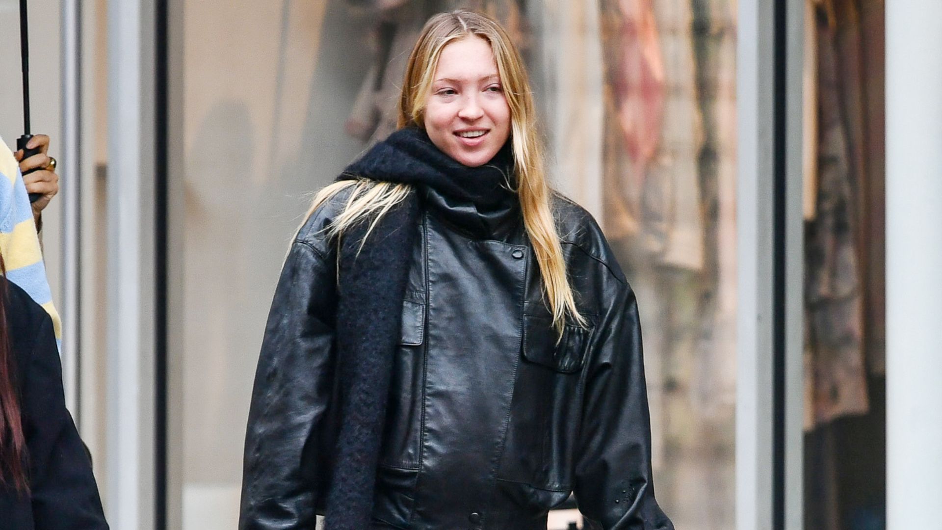 Lila Moss wears black flared trousers with a black leather jacket and heeled boots.