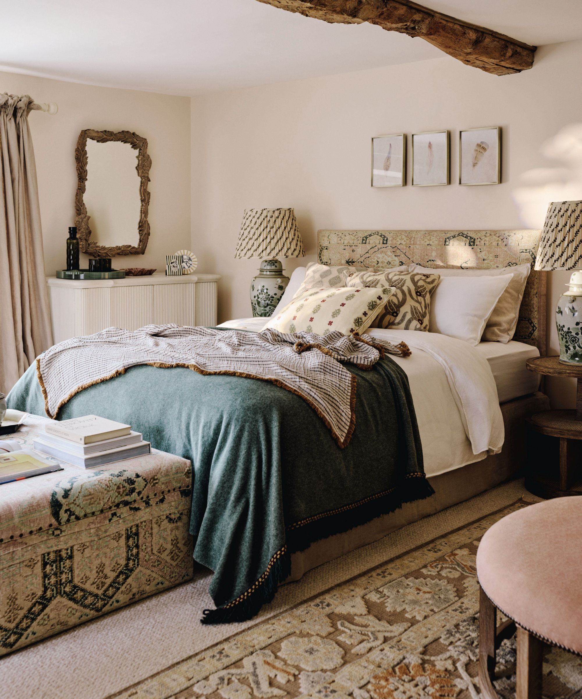 A warm, traditional bedroom with soft cream walls and a rustic wooden ceiling beam overhead. The bed is layered with white linens, patterned cushions, and a deep green throw blanket. Matching ceramic lamps sit on wooden bedside tables, while framed artwork hangs above a textured headboard. At the foot of the bed, a patterned upholstered bench holds stacked books, and a woven rug spreads across the floor.