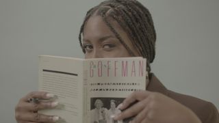 African woman holding a book in front of her face and looking over the top of it.