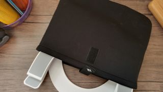 A white z-type Curve Flex laptop stand half-placed inside a neoprene bag, sitting on a brown desk