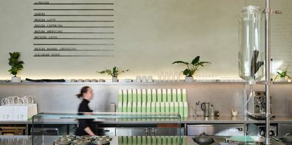 A minimalistic matcha cafe's bar features pale green plastered walls with built-in product list in black relief, chrome and glassy surfaces, a dripping water bottle, and green and cream-shaded cups.