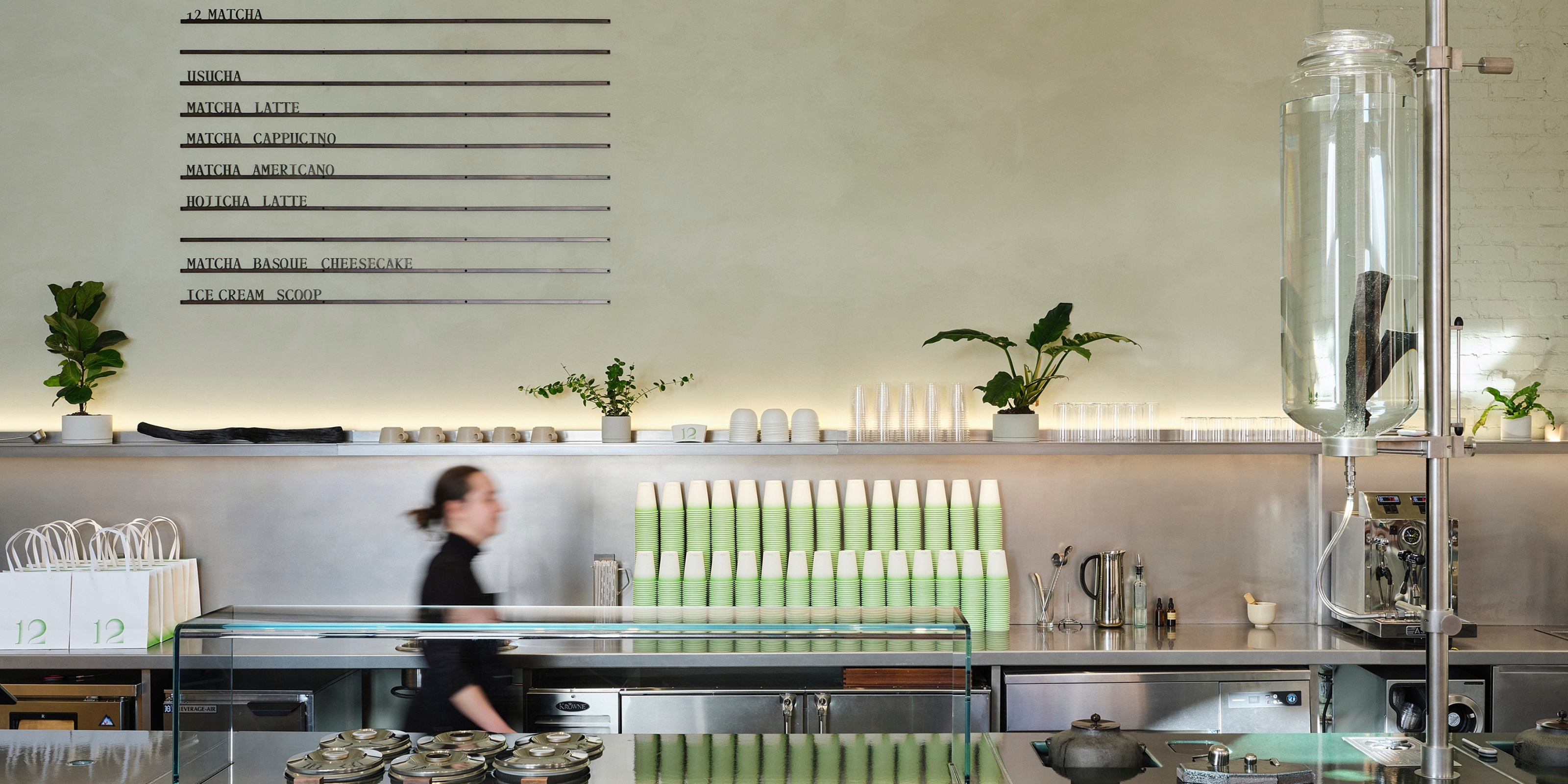 A minimalistic matcha cafe&#039;s bar features pale green plastered walls with built-in product list in black relief, chrome and glassy surfaces, a dripping water bottle, and green and cream-shaded cups.