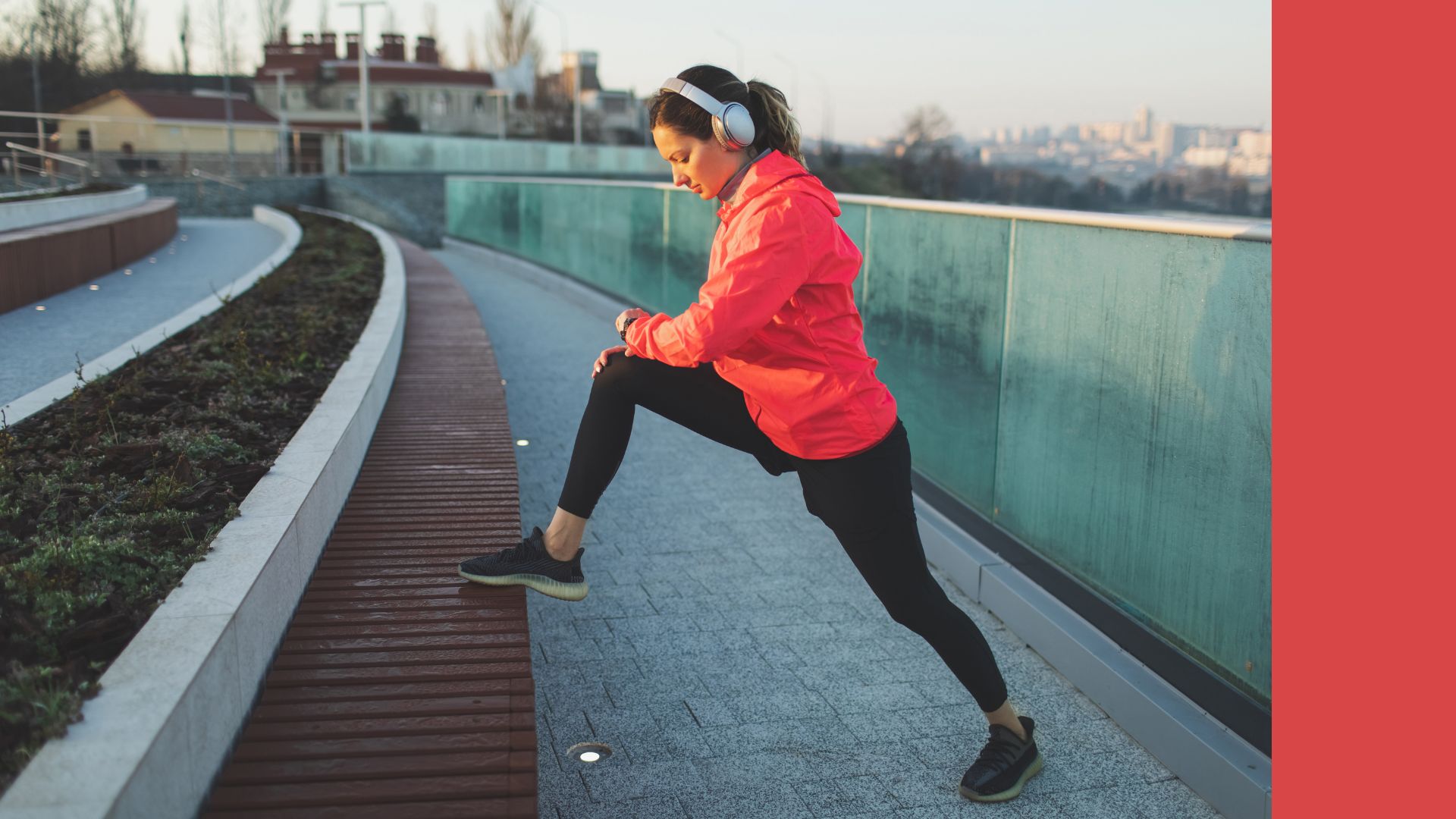Woman doing running warm-up while looking at smartwatch, wearing jacket and leggings on riverside