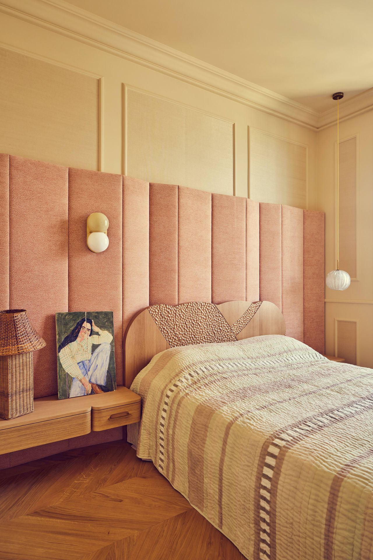 Batiik Studio-designed master bedroom furnished with mostly pastel pink and cream accents, which feel warm against the herringbone wooden floors. There is an oversized heardboard panel overhead, taking up the better portion of the wall.