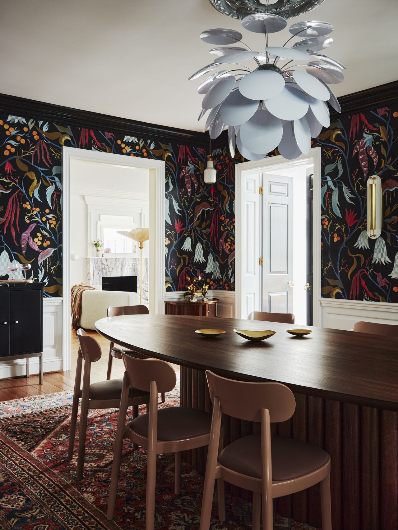 A dining room with wallpaper on the walls, wooden dining table and a chandelier above