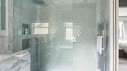 A marble and glass tiled shower with a shower niche, large glass screen, silver radiator and marble sink