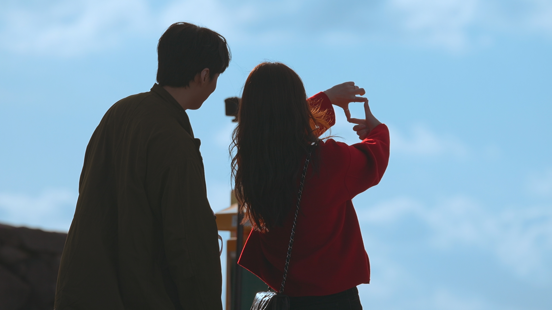 A man and woman are seen from the back as they stand under a blue sky. The woman is making a square with her fingers, in the still from Netflix's 'Better Late Than Single' season 2.