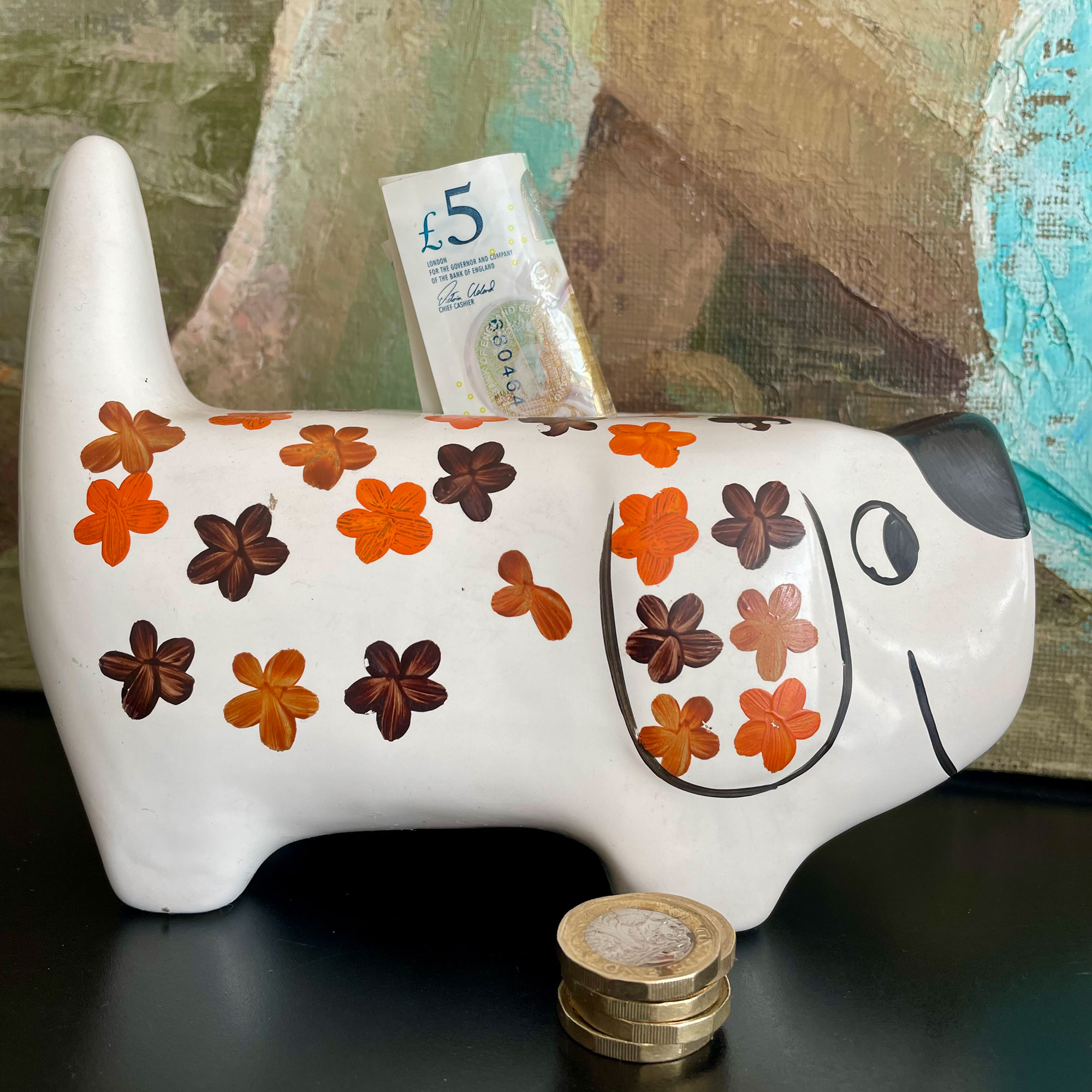 Vintage piggy bank with orange and brown flowers with pound coins by it and five pound note sticking into it