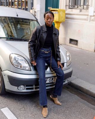 Sylvie Mus wears a black leather jacket, a black turtleneck sweater, blue jeans, light brown mules and a gold chain belt.