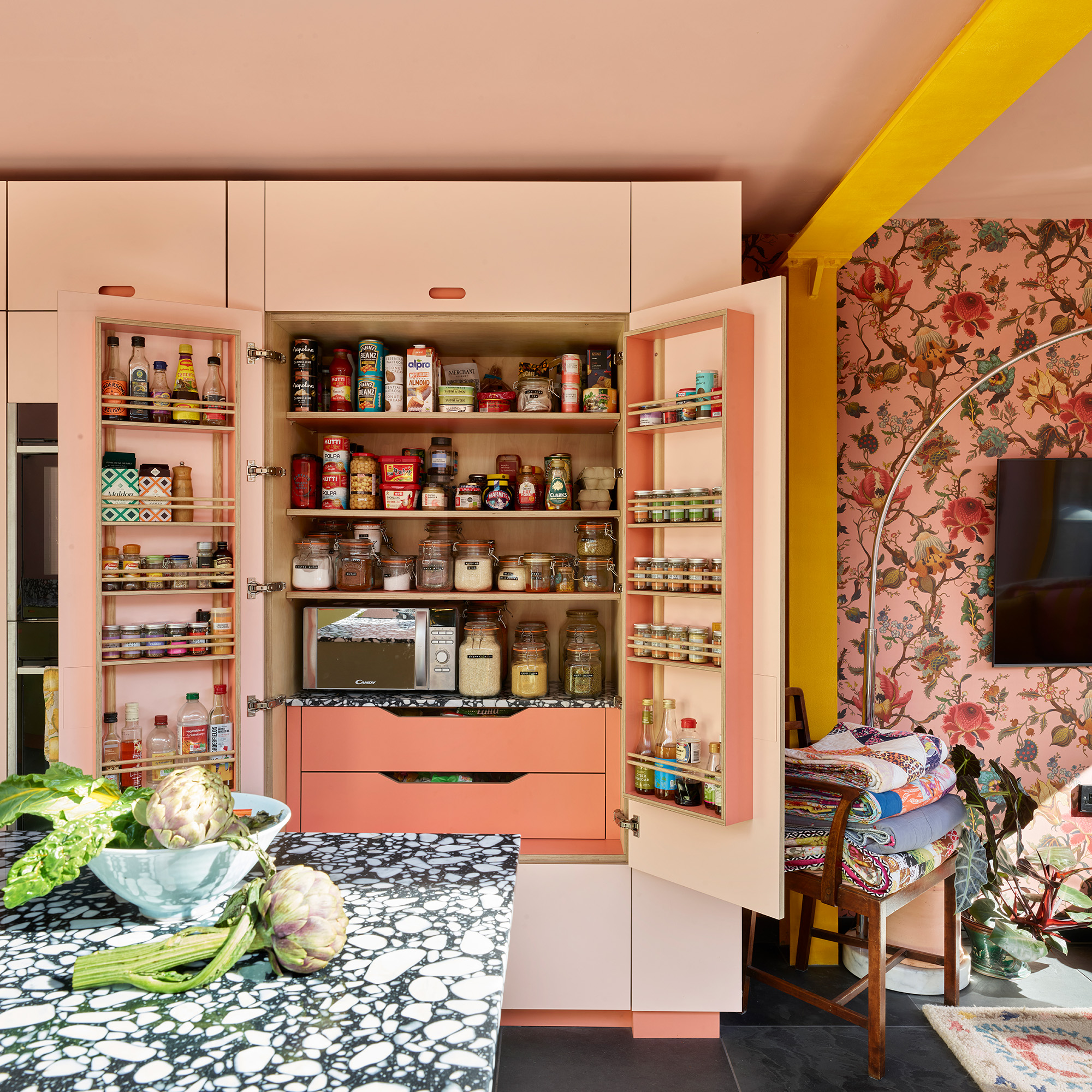 open plan kitchen with pink walls and floral pink wallpaper large double door pantry cupboard integrated into a run of full height cabinetry black and white speckled worktop in foreground