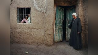 Person in traditional clothing entering a weathered door in a mud-brick building, child sitting in a window to the left