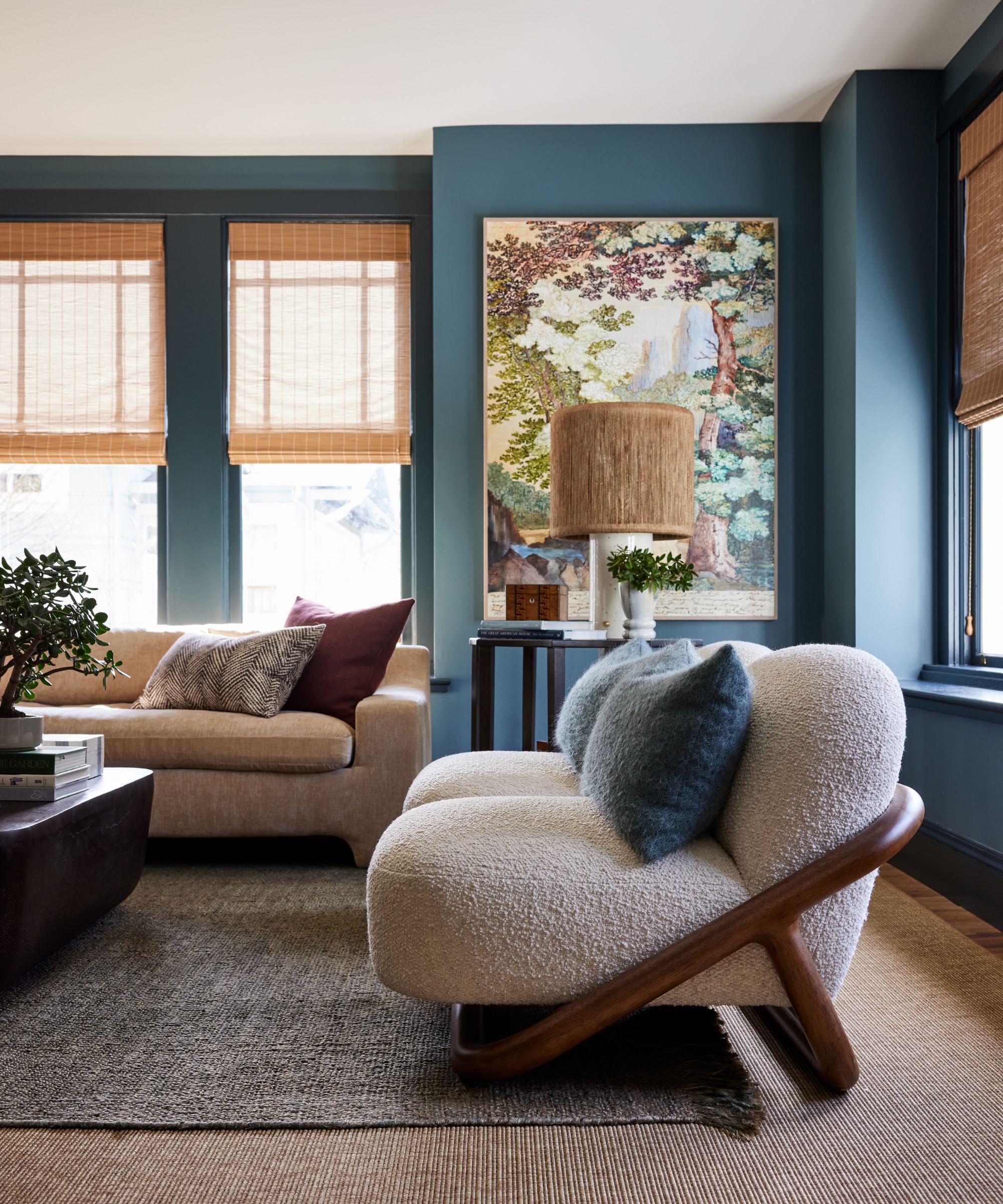 A modern living room with muted blue walls, windows with bamboo Roman blinds, a neutral carpet with a darker layered rug, a beige sofa, and two cream boucle armchairs. Soft blue and burgundy accent cushions, a floral-motif artwork on the wall, and a woven lampshade.
