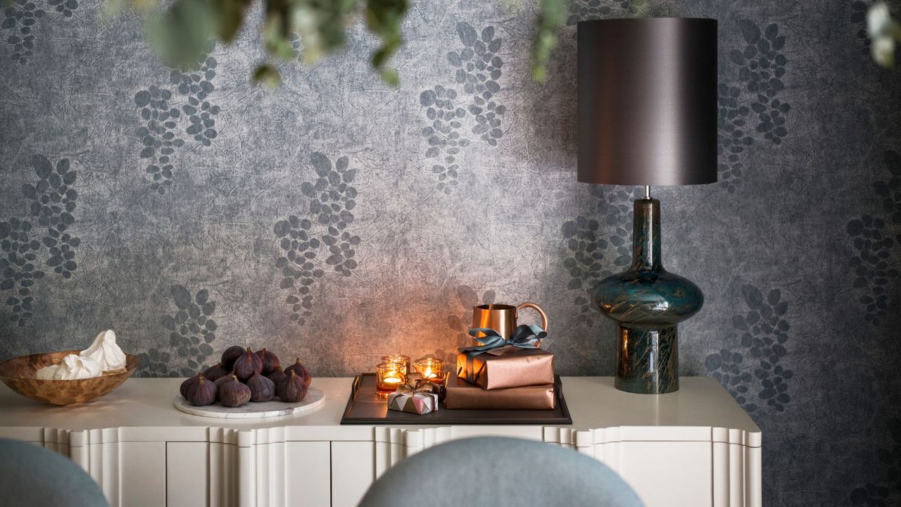 A white side table with a wooden bowl of meringues, a white plate of figs, and a tray with tealight candles, presents wrapped in bronze paper, a small bronze jug, and a glass lamp with a grey lampshade. The walls are wallpapered with silver paper with grapes on.