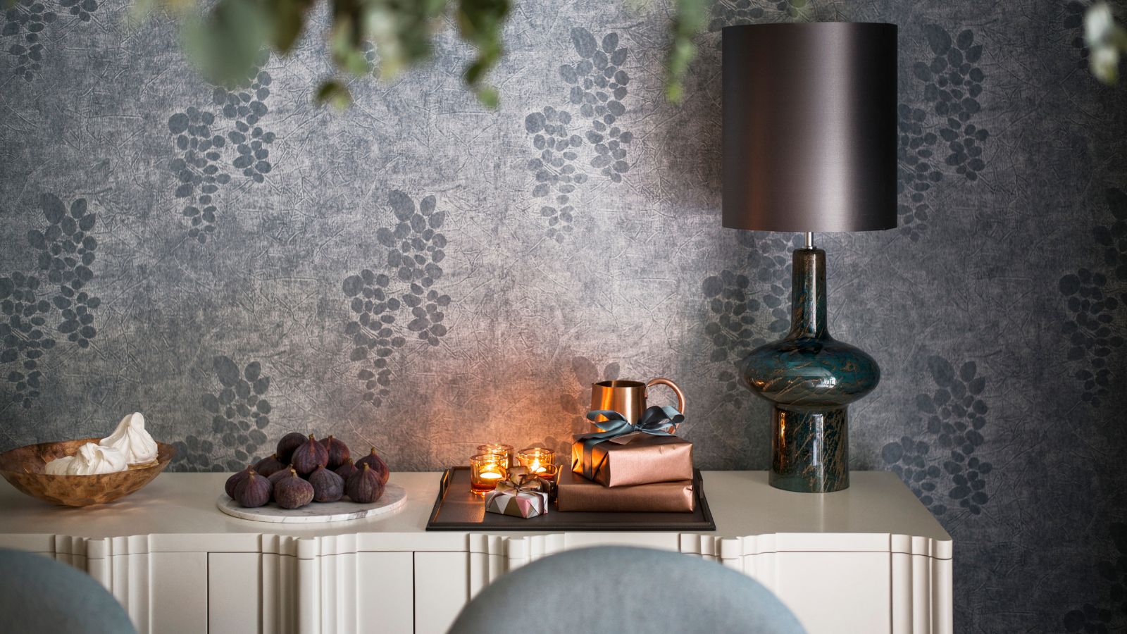 A white side table with a wooden bowl of meringues, a white plate of figs, and a tray with tealight candles, presents wrapped in bronze paper, a small bronze jug, and a glass lamp with a grey lampshade. The walls are wallpapered with silver paper with grapes on.