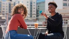 Photo of a young woman and a young man having iced coffee outside. The man is holding up a credit card.