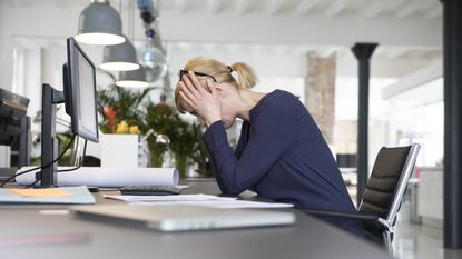 A woman sits at her desk in an office with her head down and hands over her ears.
