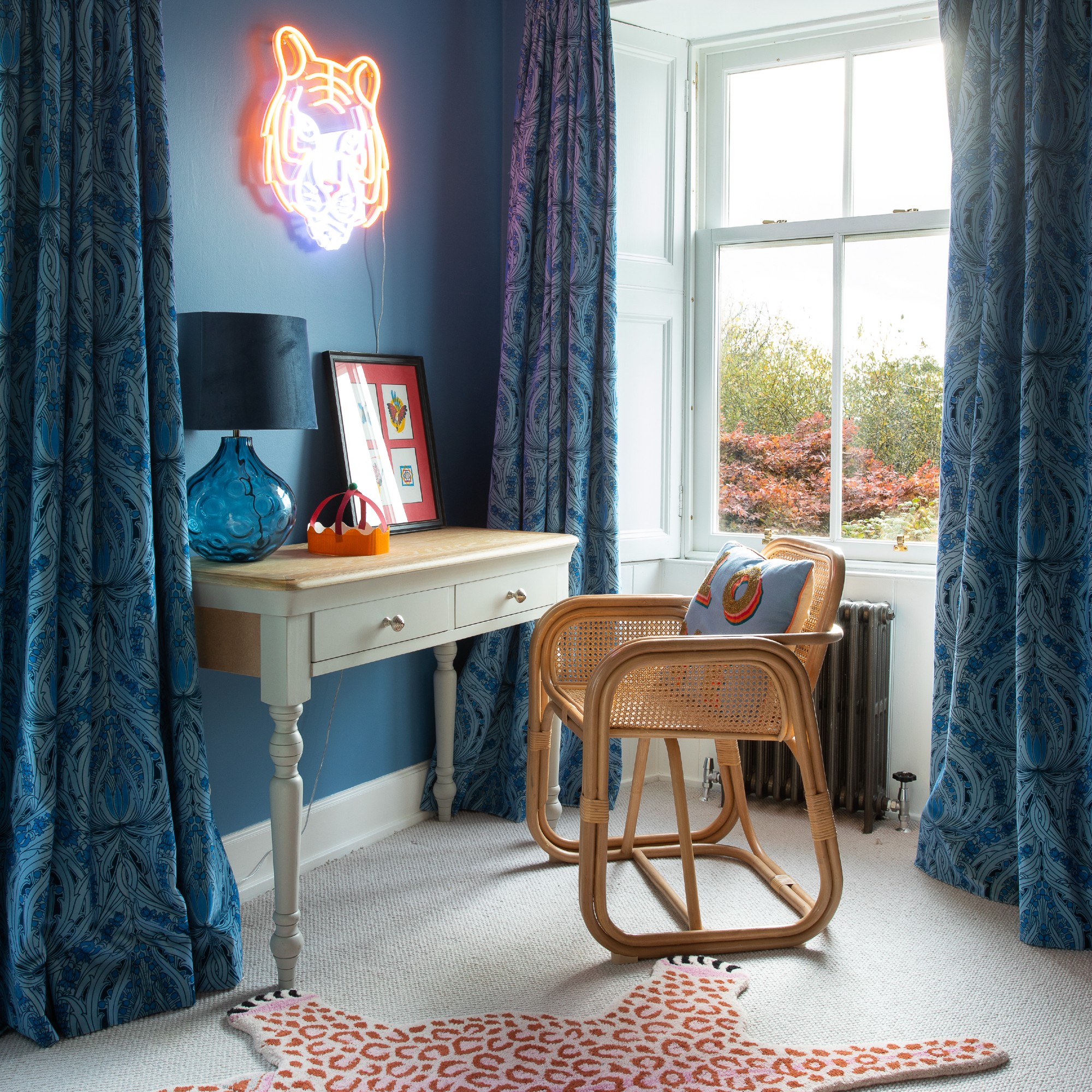 A blue-painted home office with matching patterned curtains, a tiger neon sign above the desk and a blue table lamp