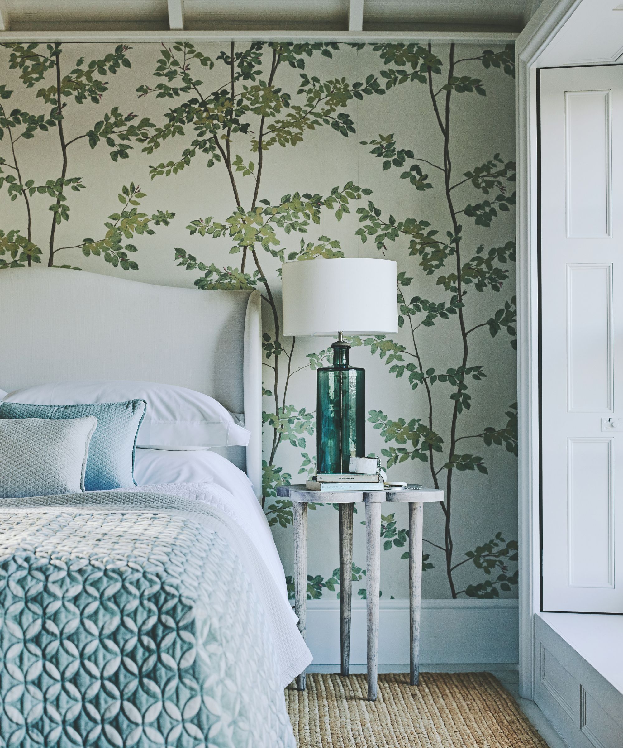 A wallpapered bedroom with green trees on it, a large white bed with blue bedding, wooden bedside table with green blue glass lamp with white lampshade, and large white shutters seen to the right. The carpet is textured and woven.