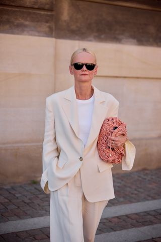 A woman wearing an ivory oversize blazer with matching trousers, a knit clutch, and black sunglasses.