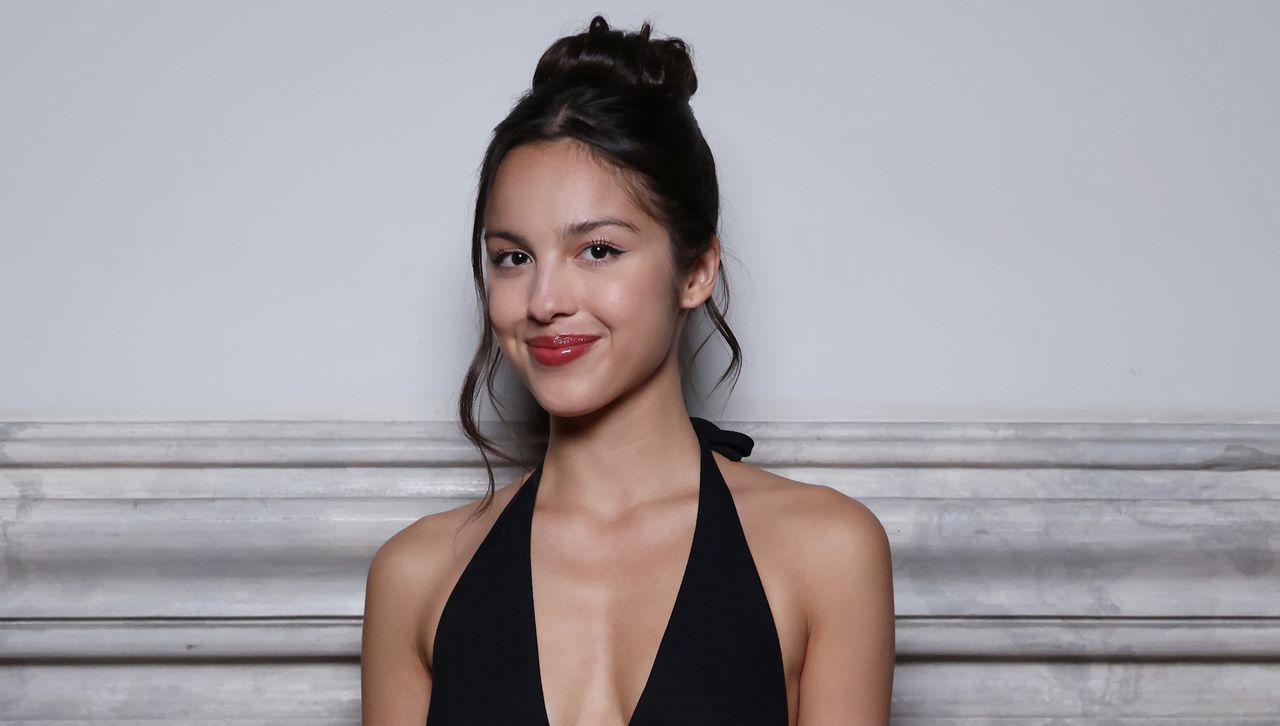 Olivia Rodrigo wears a halter dress with a high bun while standing in front of a plain wall