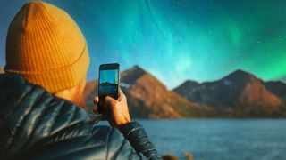 a man wearing a orange yellow hat holds up a phone to take a photo of northern lights dancing in the sky as ribbons of green and blue light