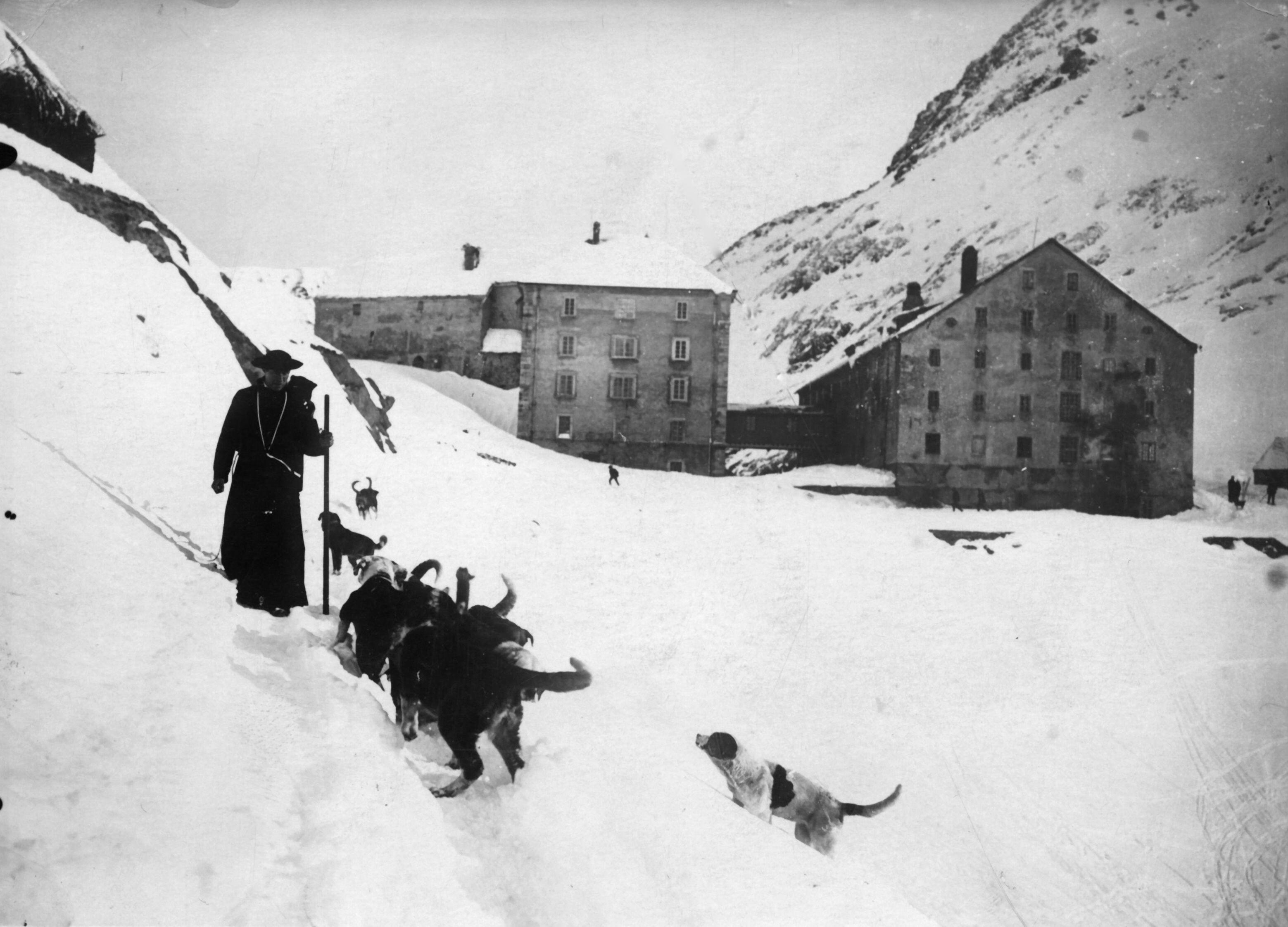 A monk walks through deep snow with a group of St Bernard dogs following behind him; large stone hospice buildings sit in the snowy mountain landscape in the background.