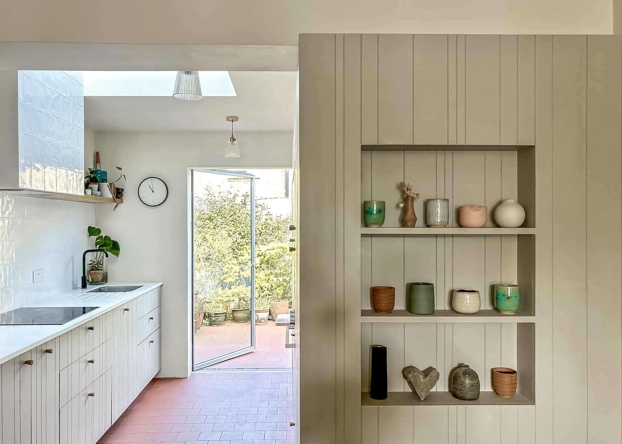 A view of the kitchen with built in shelves to the right, in a sage/avocado green colour. To the left is a view through to the garden with the large glass door at the back, a large skylight above the induction hob and the sink in the corner.