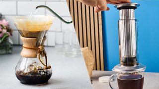 an hourglass coffee maker by chemex is brewing black coffee on the left side and an aeropress constructed of glass and metal is brewing coffee on the right side