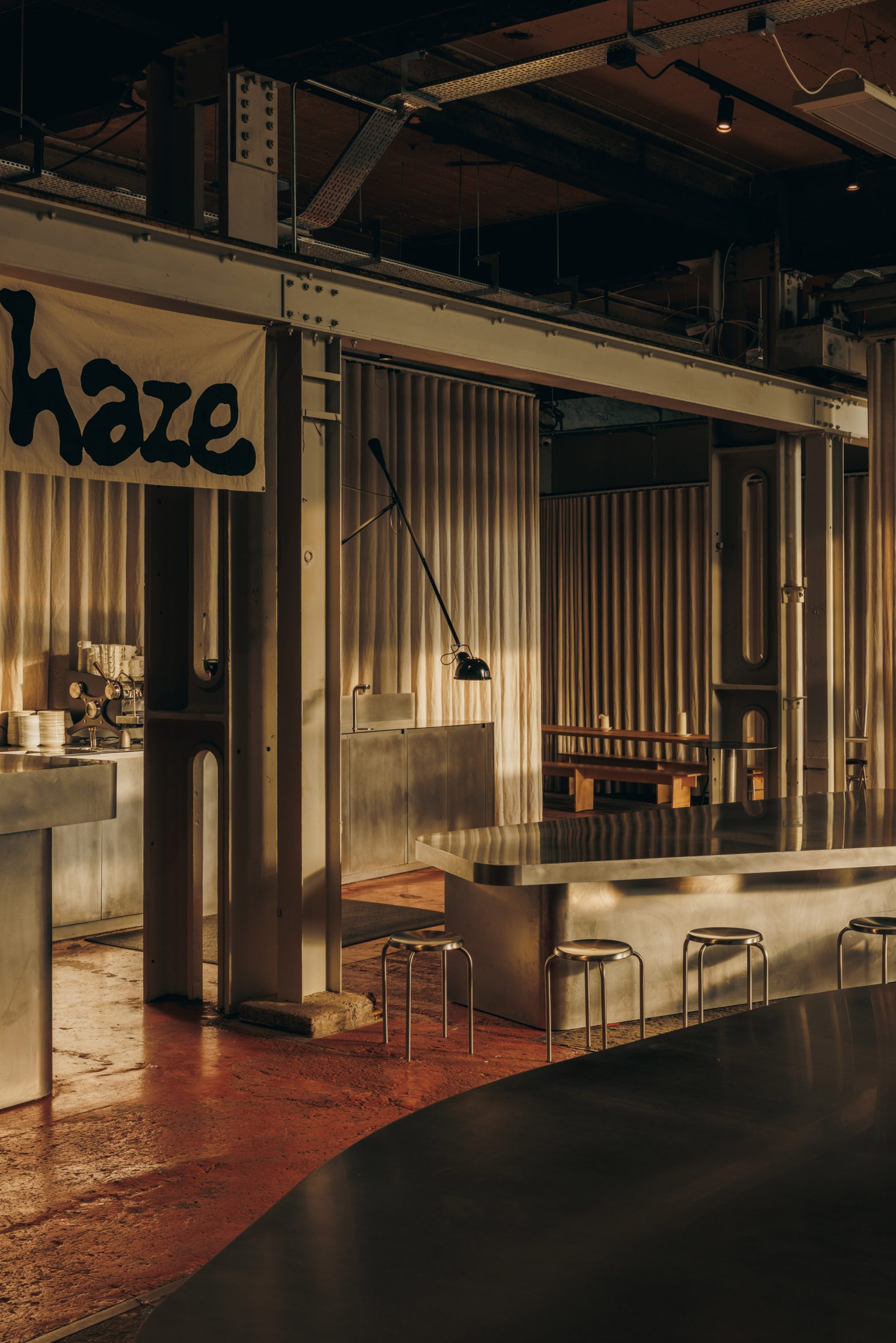 A former warehouse now converted into a restaurant features matte chrome countertops, beige curtains, chrome stools, and raw red cement floors, with metallic architectural elements exposed.
