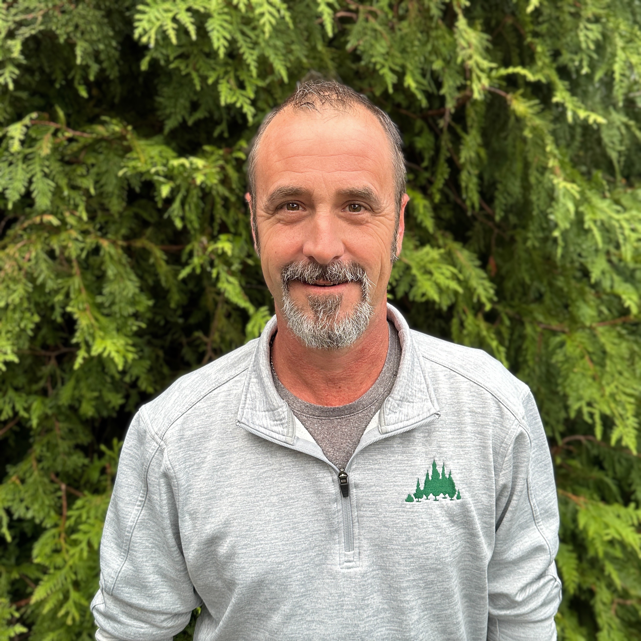 a middle aged white man with a beard smiling to camera in front of evergreen trees