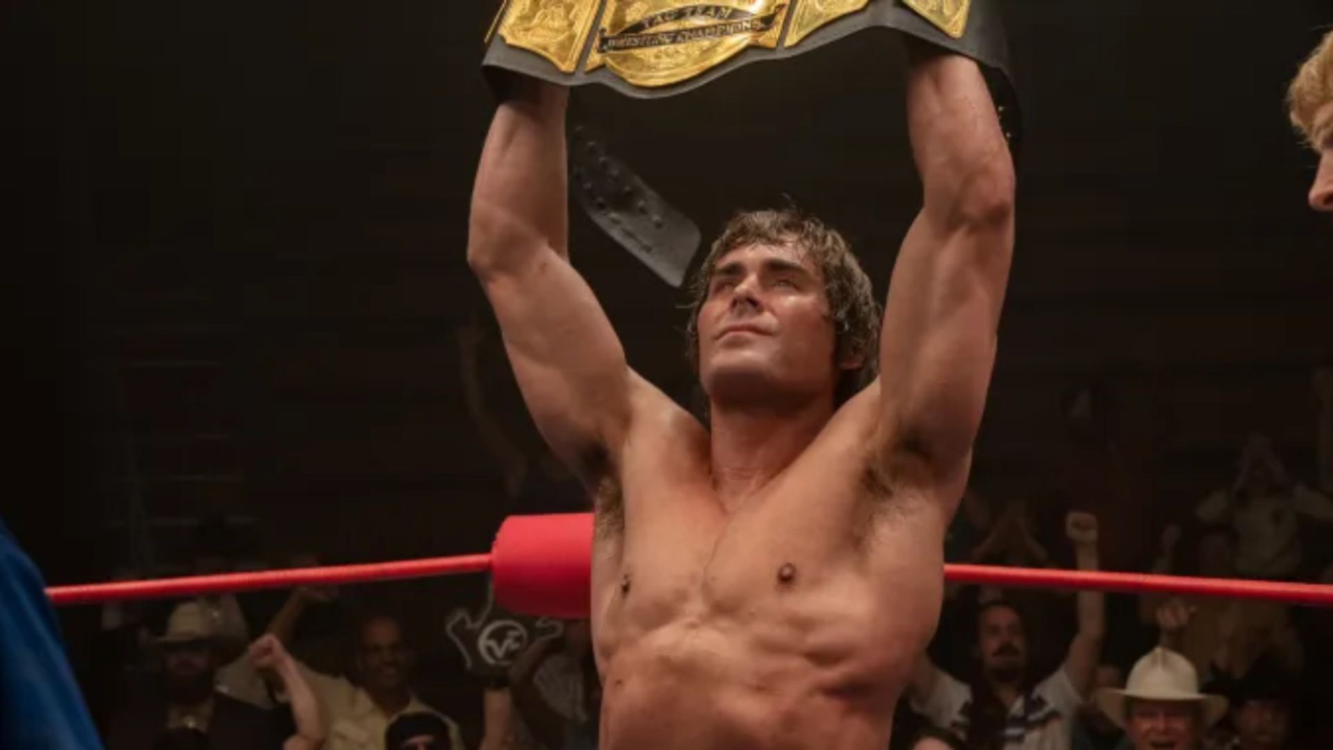 an image of Zac Efron in character int he film The Iron Claw. He is holding the winning cummerbund above his head after winning a match