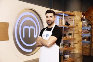 Craig Doyle stands in the MasterChef studio in front of the giant MasterChef logo on the wall, standing with his arms folded and wearing his MasterChef apron with his name on it