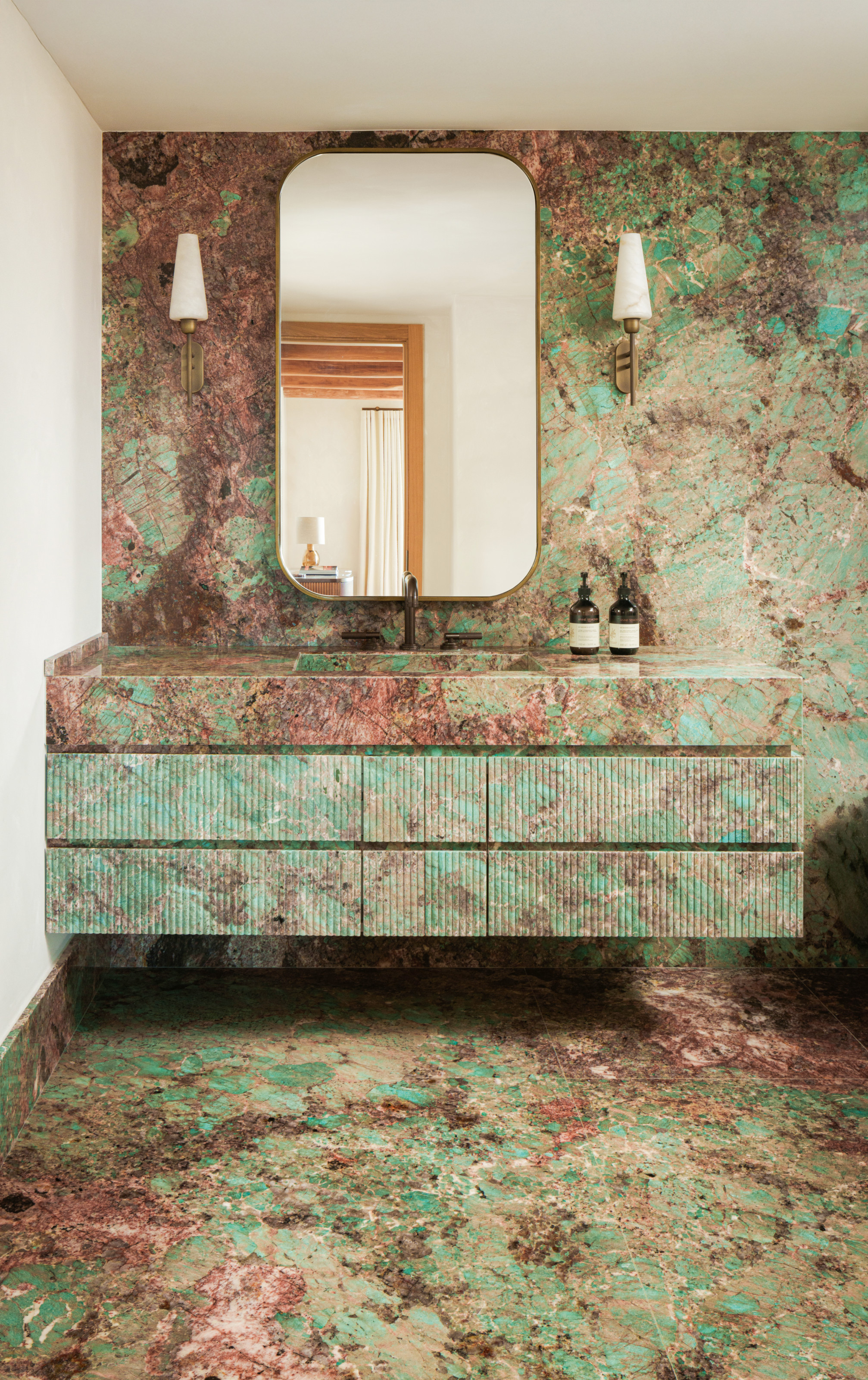 Bathroom with green and pink marble on walls, floor and vanity, with rectangular wall mirror and brass and marble wall lights