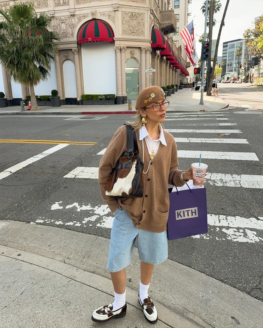 Influencer in white shirt brown cardigan, light blue denim jorts, white socks, cow print loafers and a cow print bag.