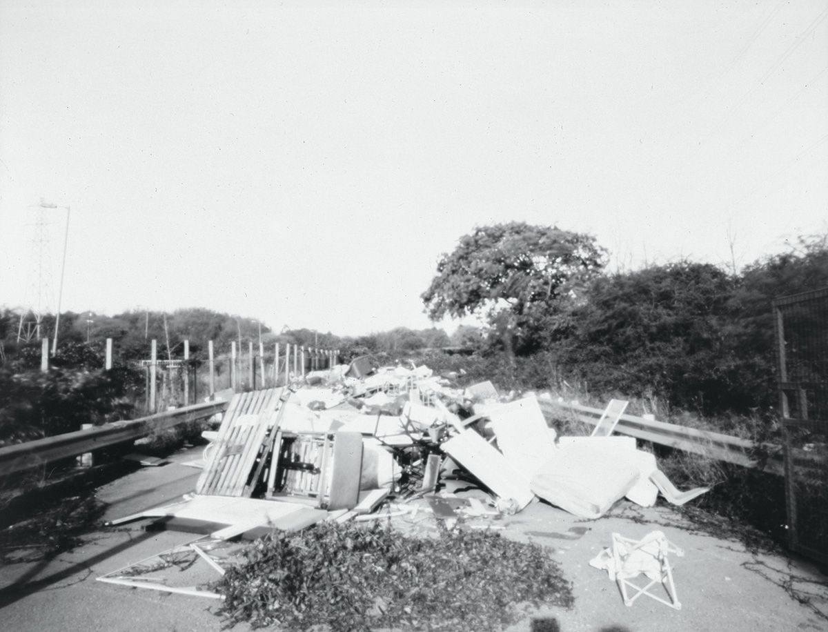 A black-and-white photograph of a large pile of dumped rubbish, including mattresses, a wooden pallet and broken furniture, blocking a tree-lined road between t