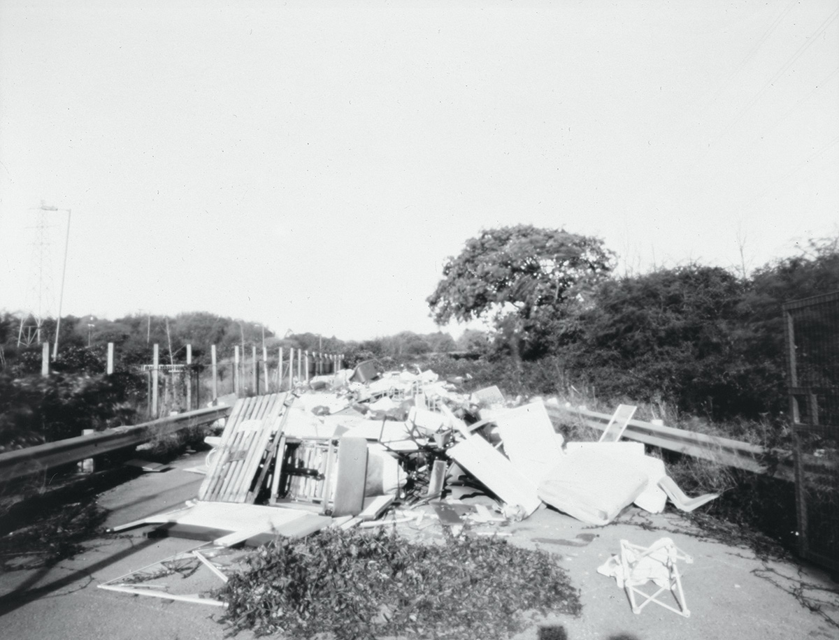 A black-and-white photograph of a large pile of dumped rubbish, including mattresses, a wooden pallet and broken furniture, blocking a tree-lined road between two crash barriers.