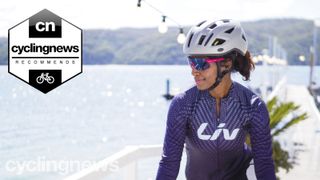 A woman cyclist wearing a purple Liv jersey, pink sunglasses and one of the best women's bike helmets stands in front of the sea