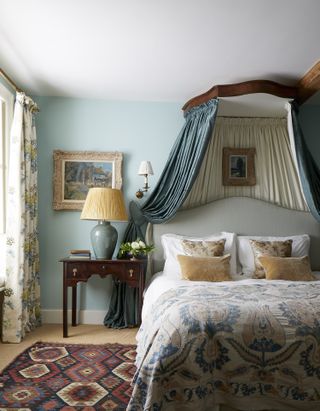 blue bedroom with white ceiling, bedding, blue fabric canopy, floral drapes, bedside, taupe and floral cushions, rug on floor