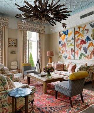 Maximalist style living room, blue painted ceiling, striking wooden pendant light, patterned curtains and pelmets, floral patterned sofa and two matching armchairs facing one another, wooden coffee table, two lounge chairs in gray checkered pattern, large red rug