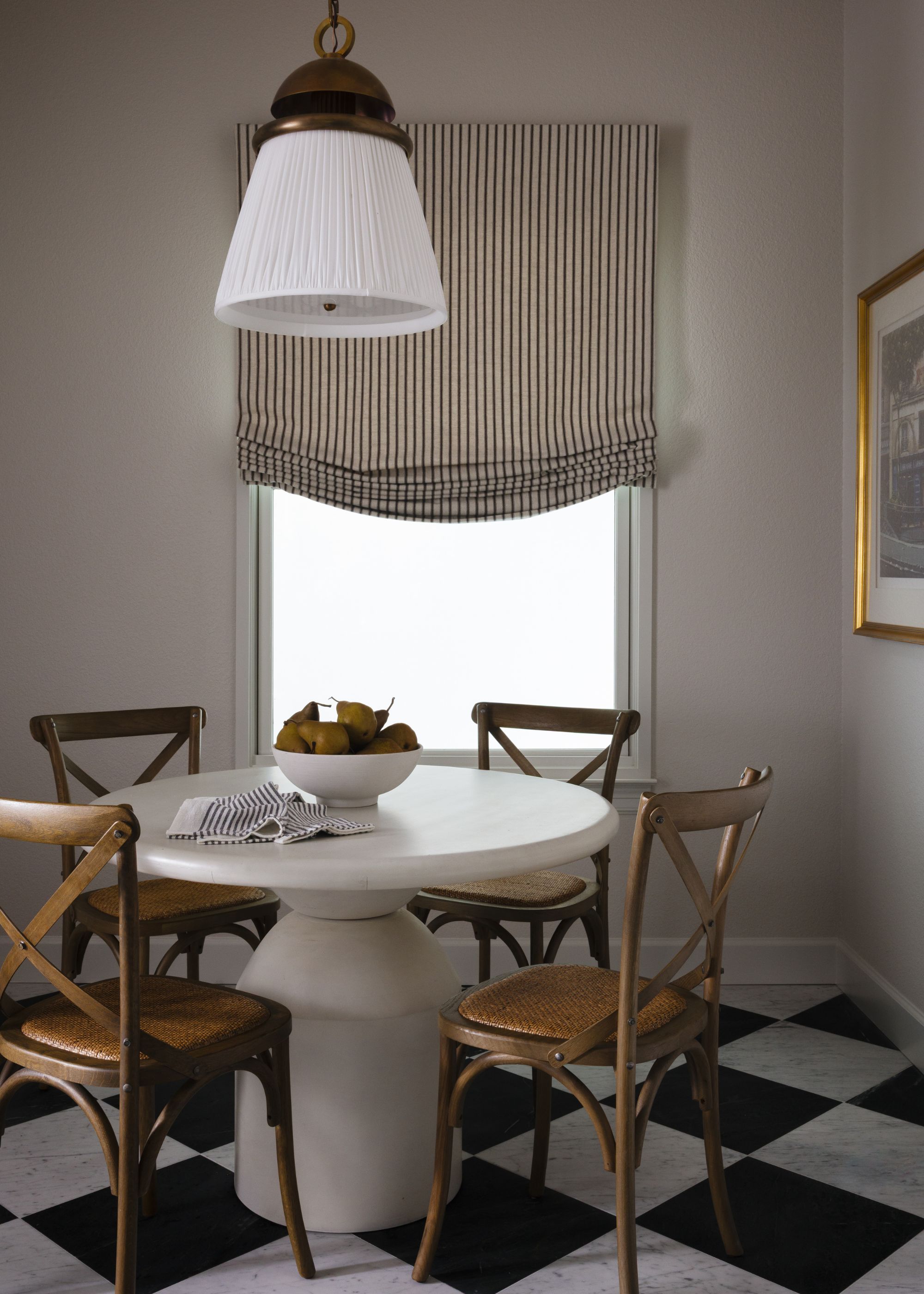 A cozy dining corner featuring a white pedestal table and four wooden cross-back chairs with cane seats. The space is accented by black-and-white checkered marble floors and a striped Roman shade.