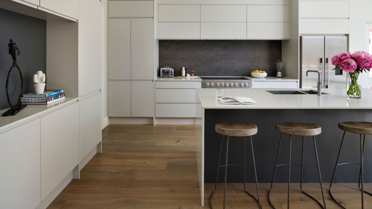 A modern white kitchen with white cabinets, wooden flooring, a dark grey backsplash, and a central white kitchen island with a dark grey space underneath the counter. At the counter are three matching metal and wood barstools, and on top of the counter is an open recipe book, glass of water, silver faucet with recessed sink, and a glass vase of large pink flowers.