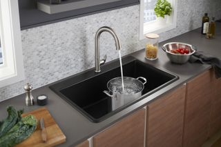 A kitchen tap with water coming out into a pot inside the sink. Featuring tiled kitchen walls, a green plant in a white vase on the white window sill. Grey kitchen top and brown cupboard.