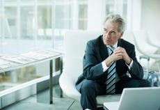 An older business man in a suit and tie sits in front of a laptop and looks to the side. He appears thoughtful or impatient.