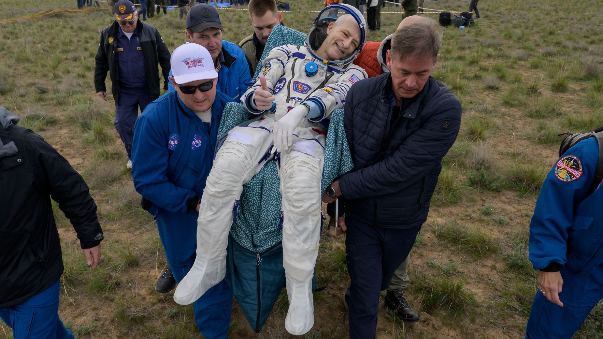 an astronaut in a white space suit smiles and gives a thumbs up as he is carried across a field in a chair immediately after landing on earth