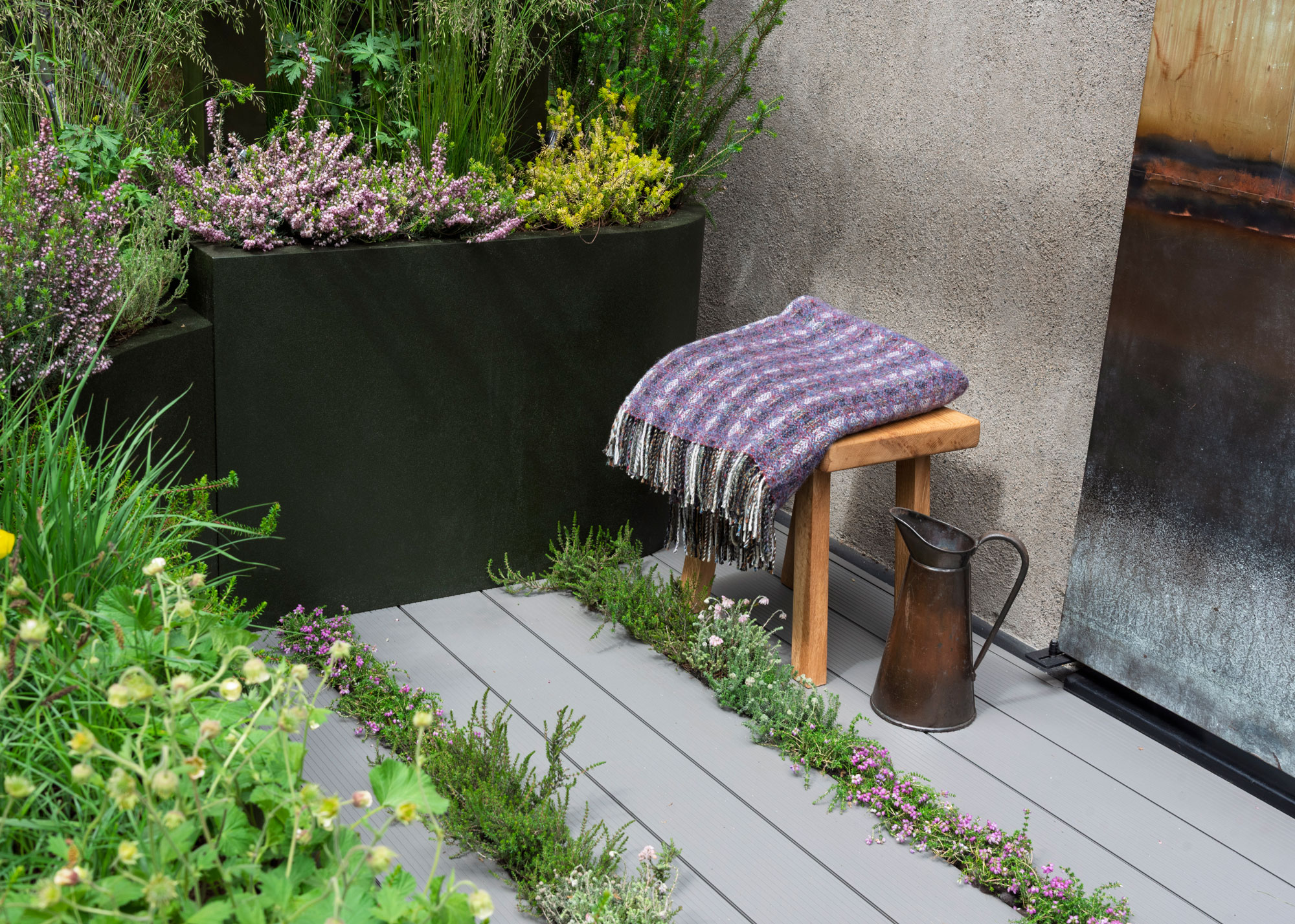 small area of deck with planting gaps, surrounded by plants, with small stool and blanket