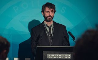 DCI Carl Morck (Matthew Goode) stands at a podium for a press conference in a scene from Netflix's "Dept. Q"