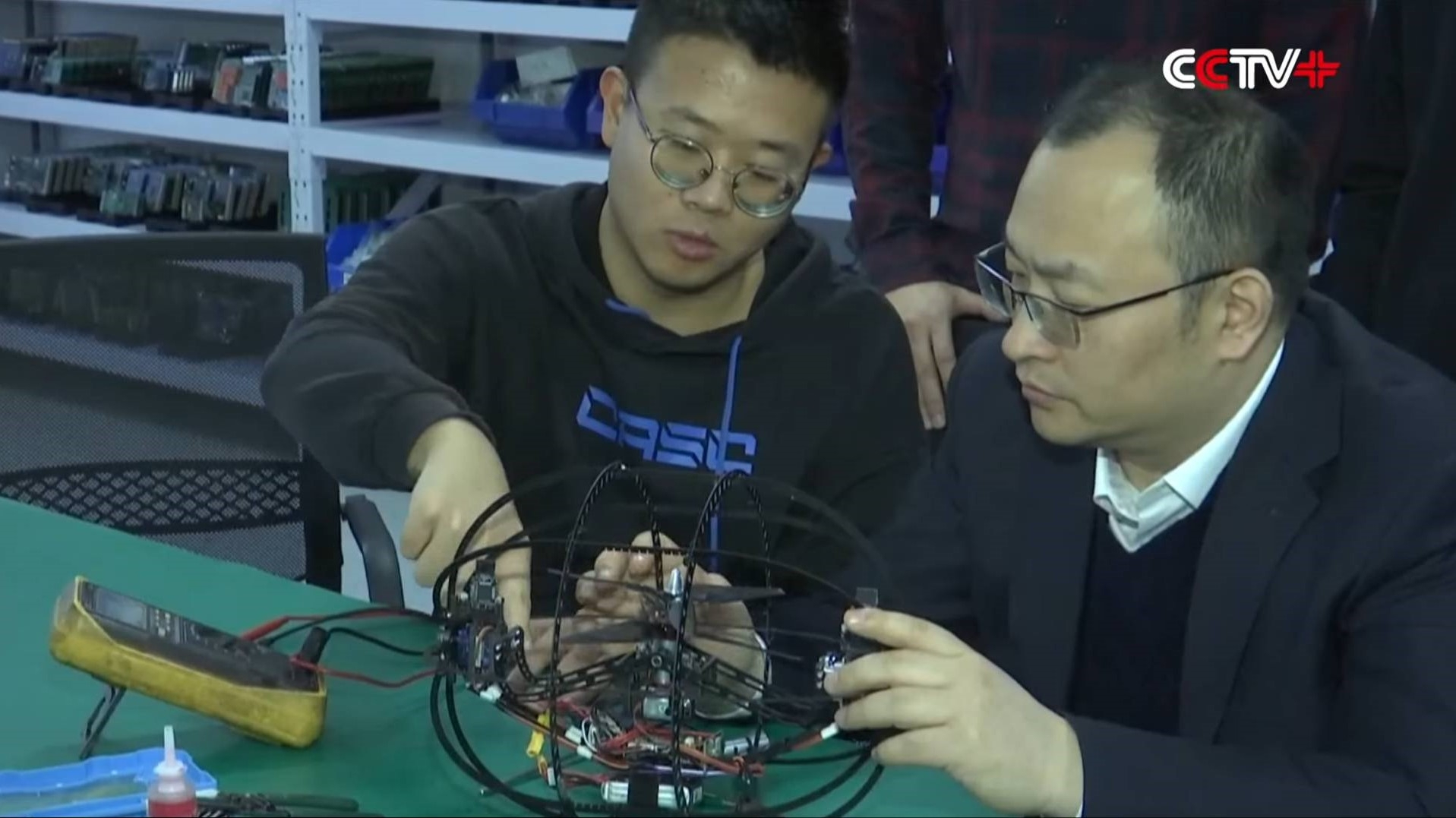 two men examine a small aerial drone while seated at a green table